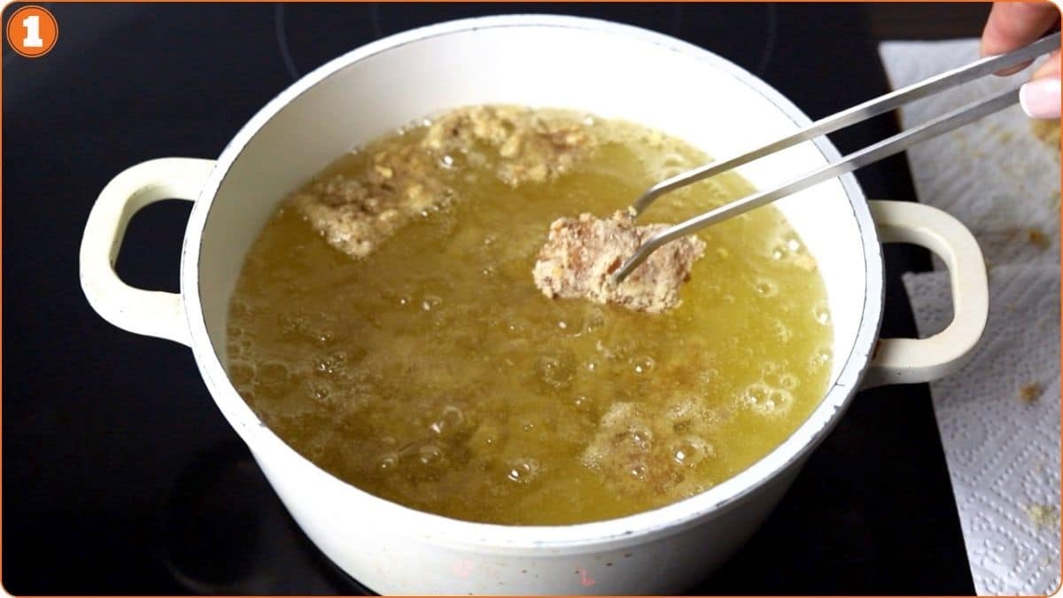 A person uses tongs to place a breaded food item into hot oil in a white pot for frying.