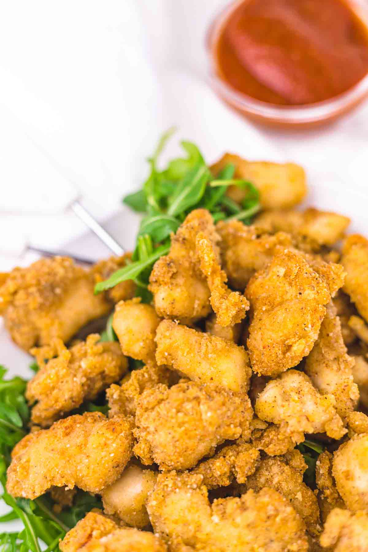 A plate of golden-brown fried chicken pieces served on a bed of greens, with a small container of red dipping sauce in the background.