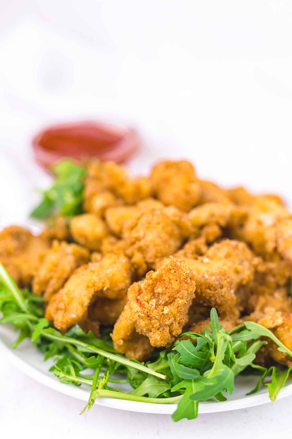 A plate of golden-brown fried chicken pieces served on a bed of fresh arugula, with a small bowl of red dipping sauce in the background.