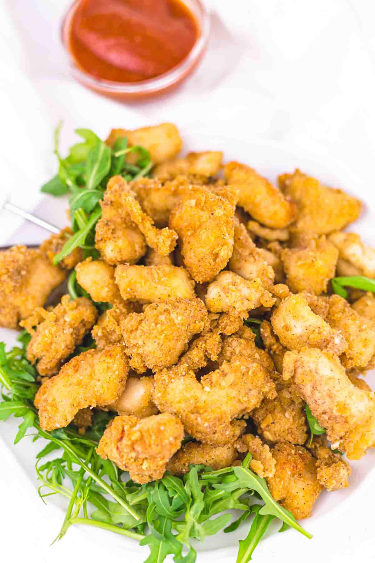A plate of crispy fried chicken pieces served on a bed of fresh arugula, with a small bowl of red dipping sauce in the background.