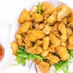 A white plate filled with crispy, breaded chicken pieces garnished with fresh green arugula, with a small bowl of dipping sauce on the side.