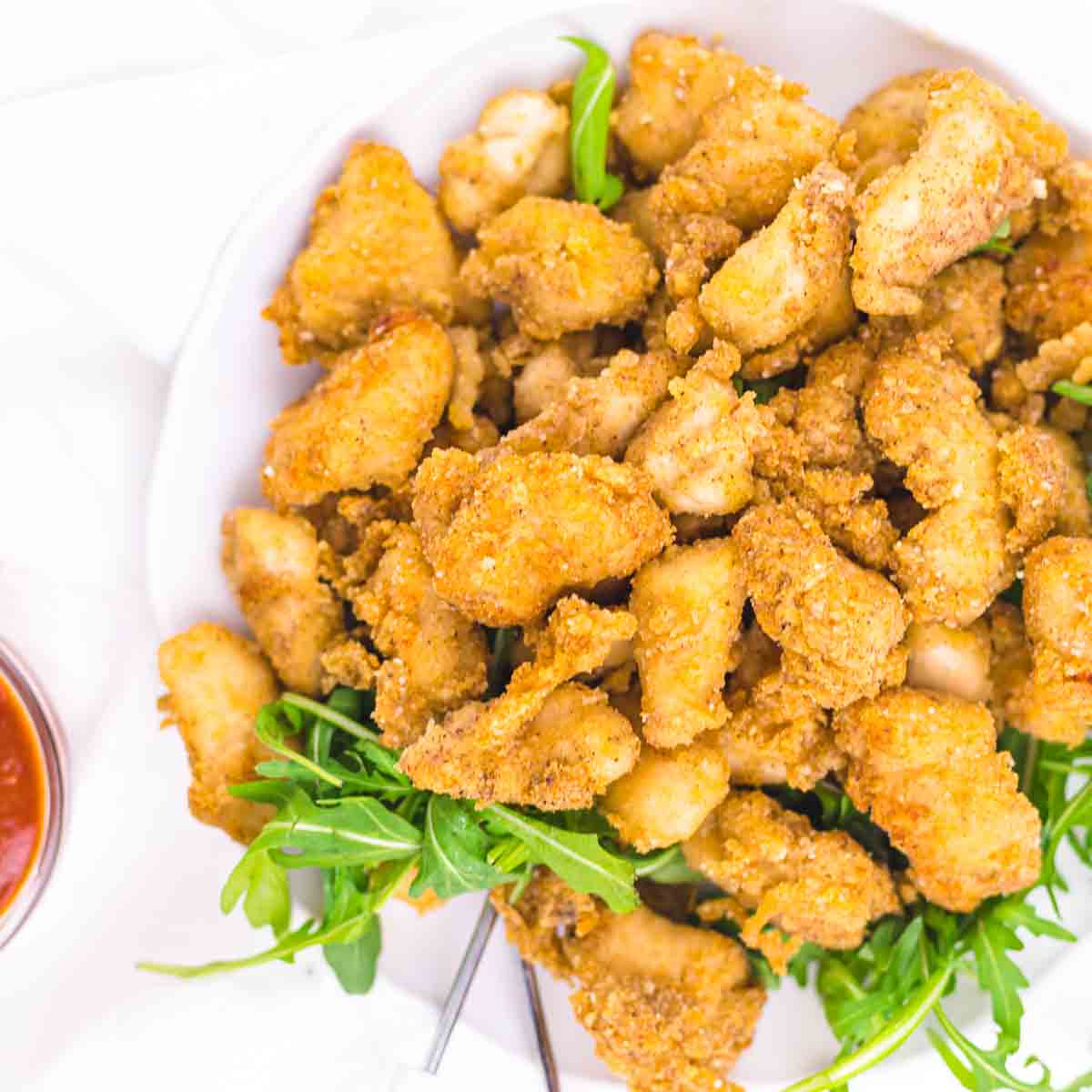 A white plate filled with crispy, breaded chicken pieces garnished with fresh green arugula, with a small bowl of dipping sauce on the side.
