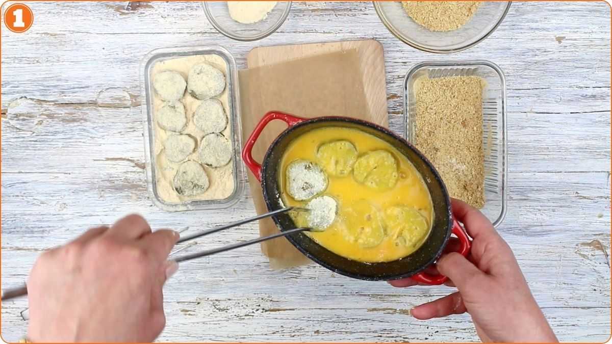 Hands dip sliced vegetables into beaten eggs using tongs, with bowls of flour and breadcrumbs arranged on a wooden surface.