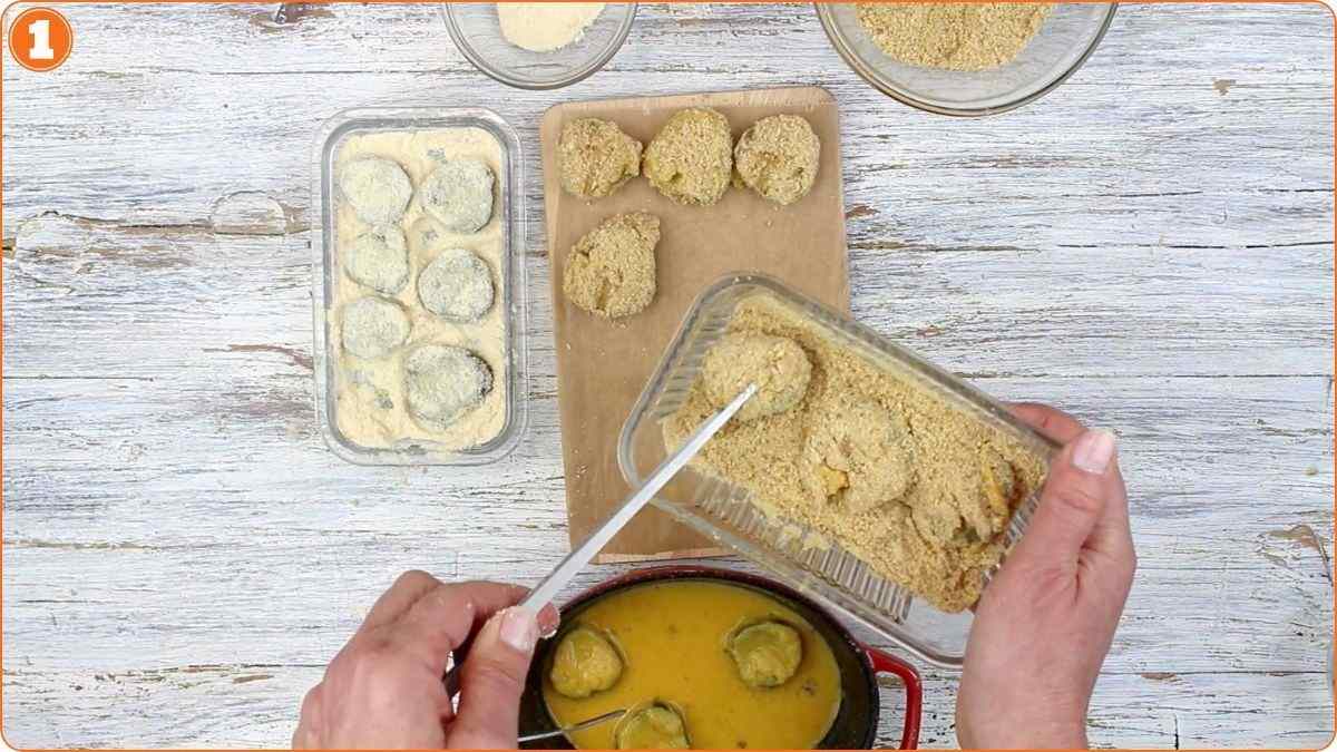 Hands coating food pieces in breadcrumbs, with bowls of egg wash and breadcrumbs, and already-breaded pieces on a tray on a white wooden surface.