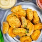 A plate of breaded and fried pickle chips is served with dipping sauces on the side.