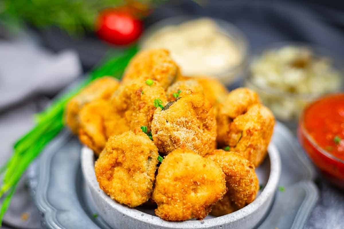 A bowl filled with breaded and fried pickle bites, garnished with chopped herbs, with dipping sauces and blurred vegetables in the background.