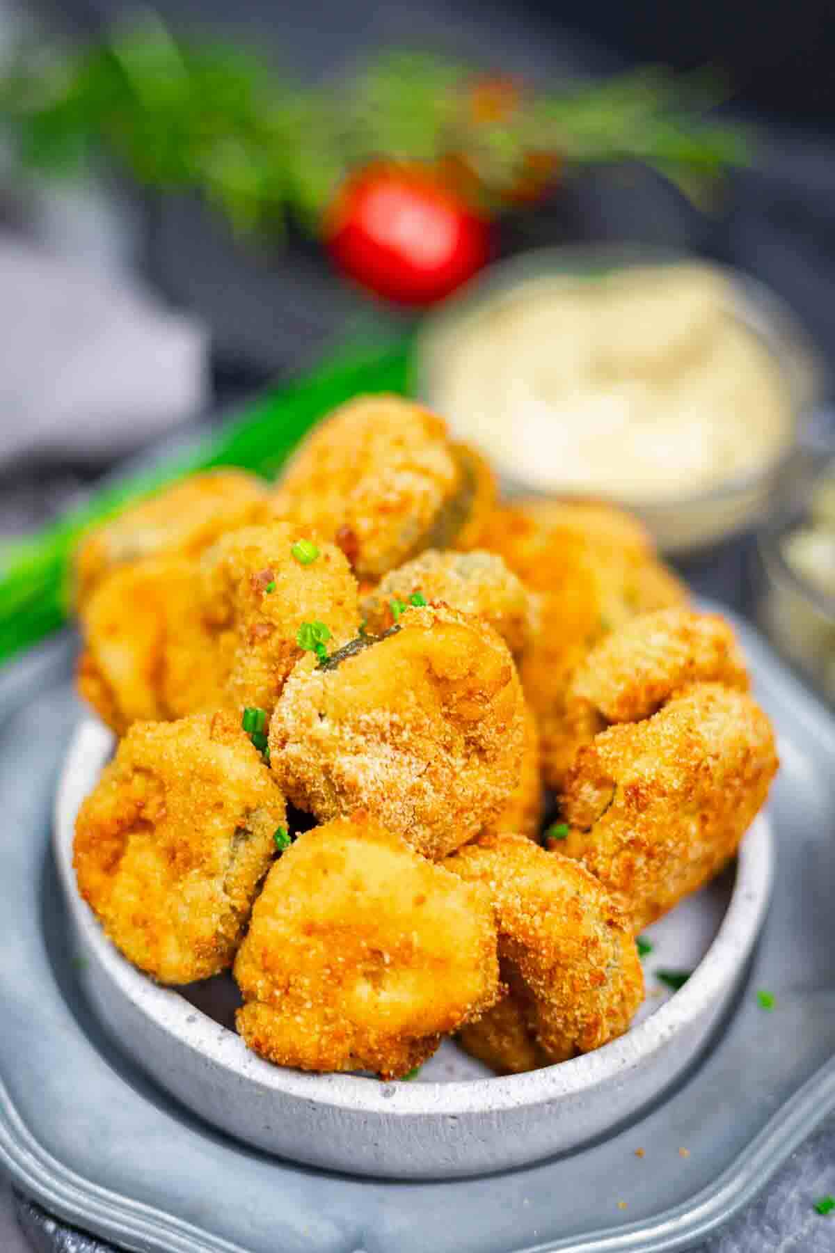 A bowl filled with breaded and fried pickle slices, garnished with chopped green herbs, with a blurred dipping sauce and vegetables in the background.