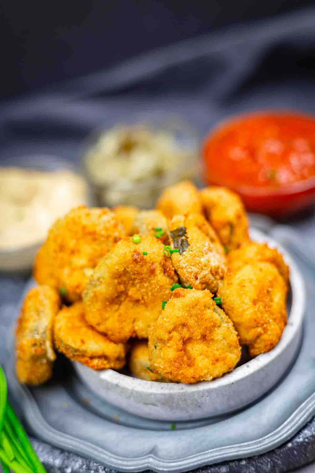 A small bowl filled with breaded, fried mushrooms, garnished with chopped herbs, with dipping sauces in the background.