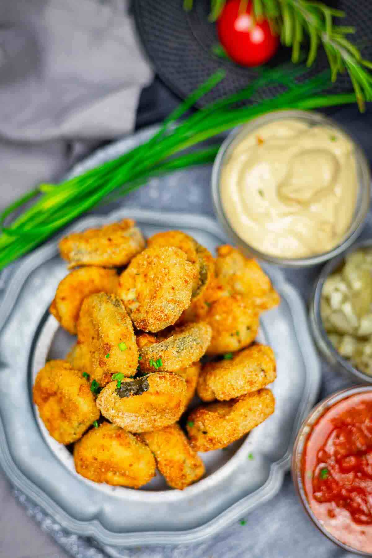A plate of breaded, fried mushroom bites served with three dipping sauces: creamy sauce, chopped pickles, and red salsa. Fresh chives and rosemary are also visible nearby.