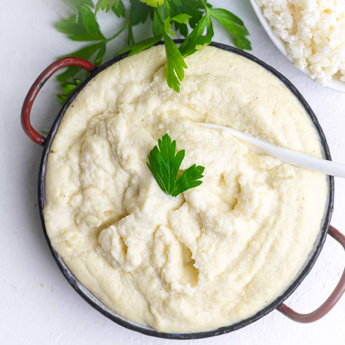 A bowl of creamy mashed cauliflower topped with a parsley leaf, with a spoon resting inside and some parsley and rice visible in the background.