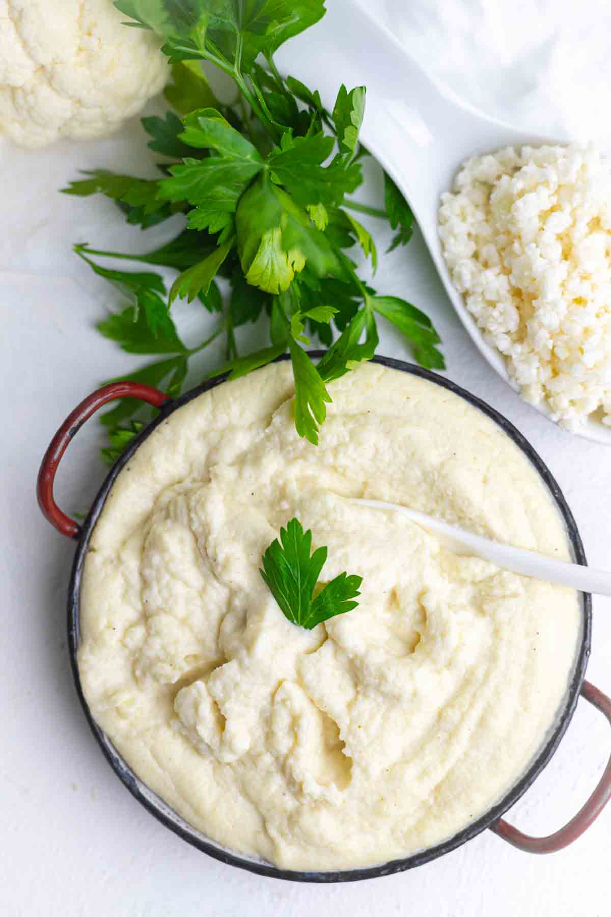 A bowl of creamy mashed cauliflower garnished with a parsley leaf, with fresh parsley and a spoonful of cottage cheese beside it on a white surface.