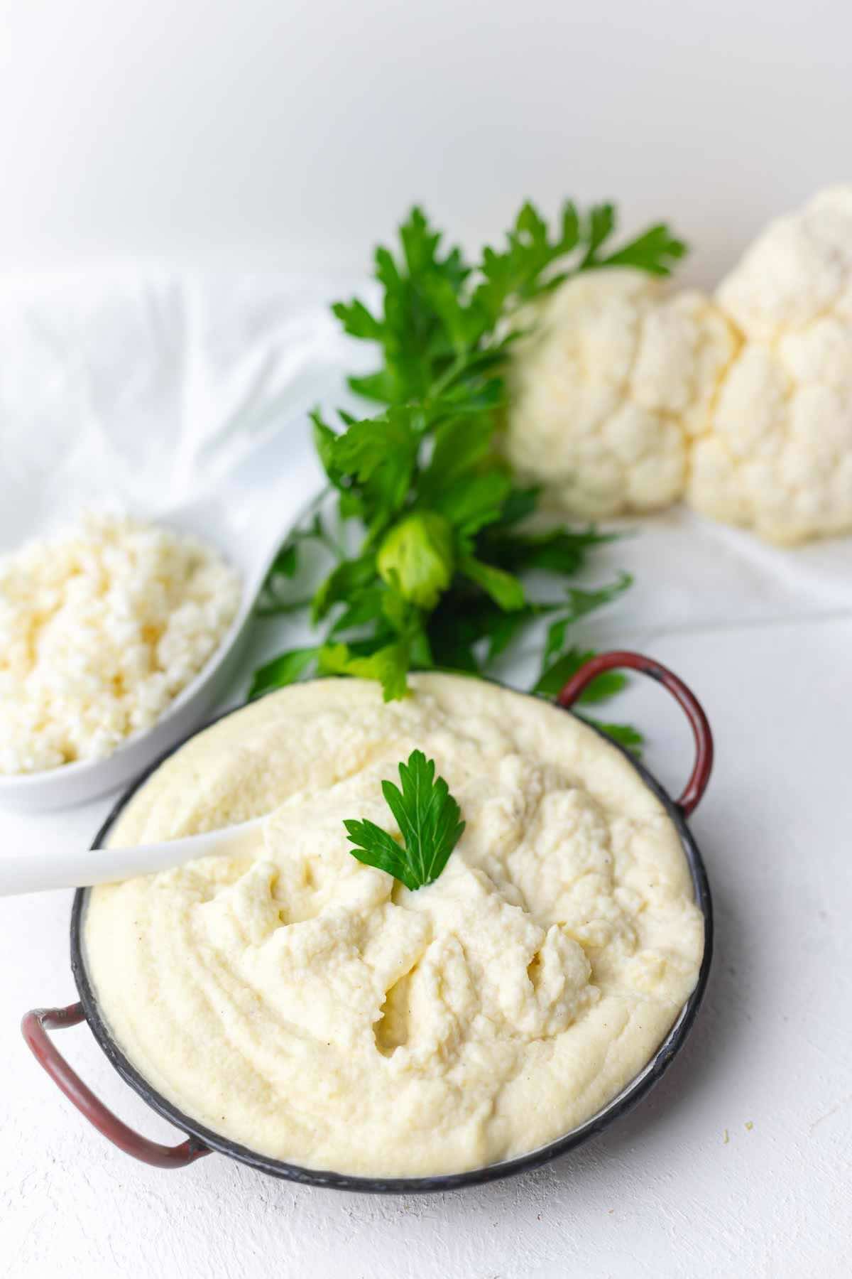 A bowl of creamy cauliflower mash garnished with a parsley leaf, with raw cauliflower florets and parsley in the background.