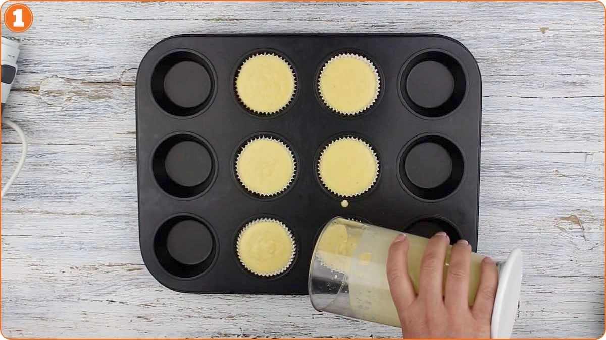 A hand pours cupcake batter into paper liners arranged in a muffin tray on a wooden surface.