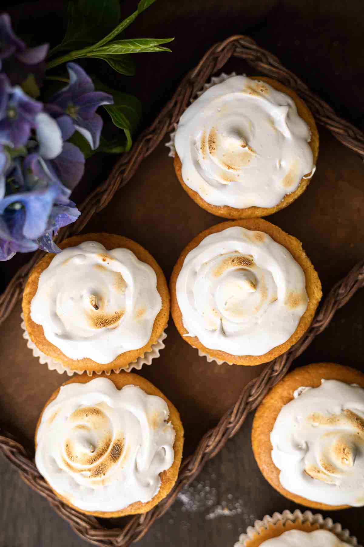 A wicker tray holding five cupcakes topped with swirled, toasted meringue frosting, next to a bouquet of purple flowers.