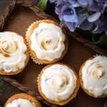 Five cupcakes with swirled white frosting are arranged on a brown tray next to a bouquet of purple flowers.