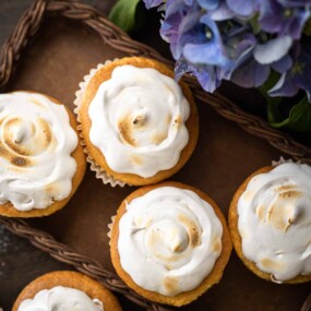 Five cupcakes with swirled white frosting are arranged on a brown tray next to a bouquet of purple flowers.