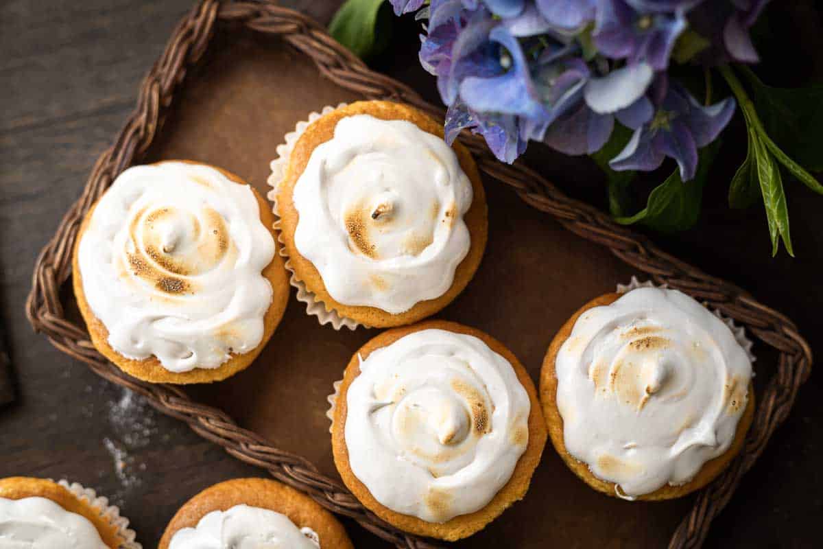Five cupcakes with toasted meringue frosting arranged on a wicker tray beside purple flowers on a wooden surface.