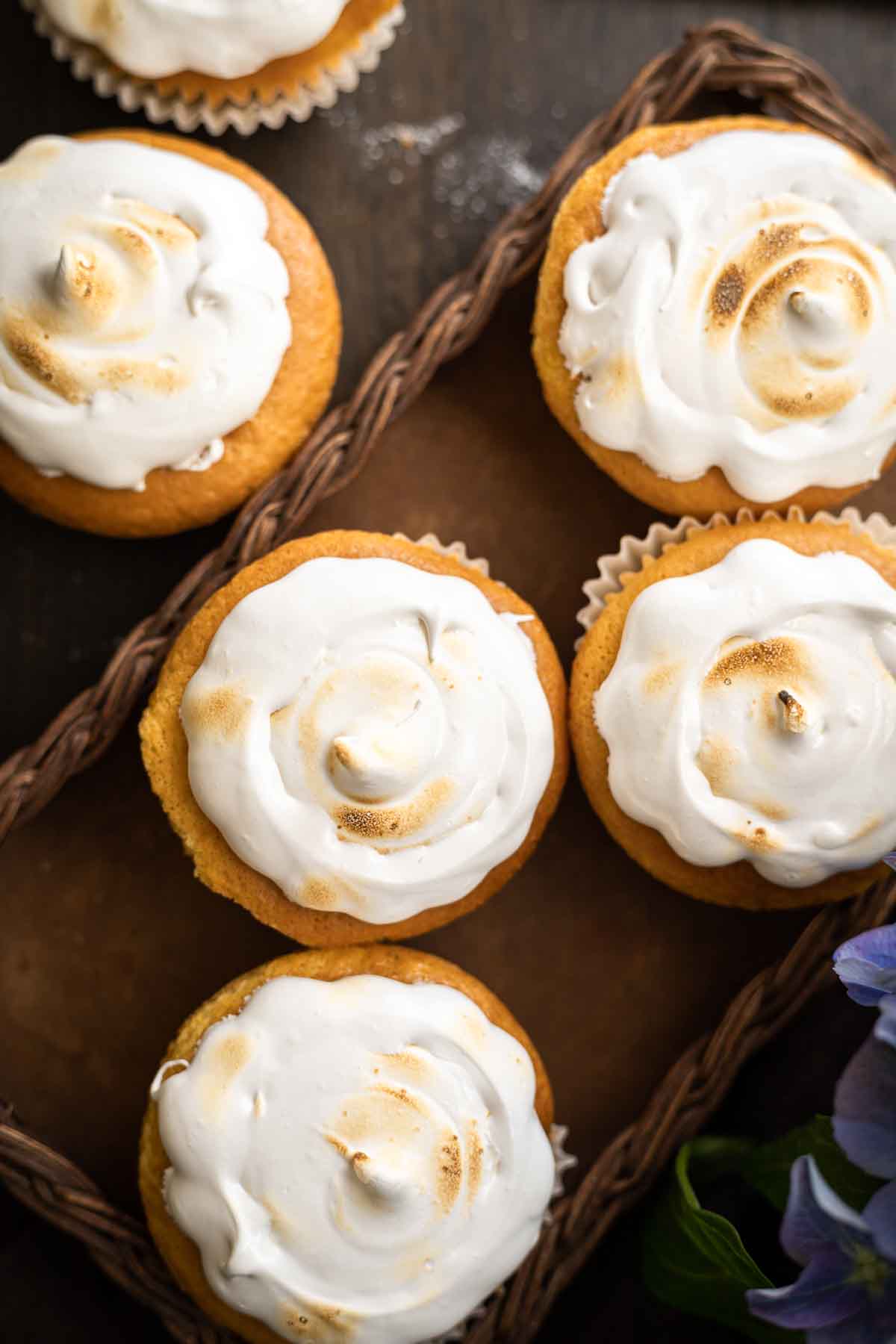 Six cupcakes with golden brown cake bases and swirled toasted meringue frosting arranged on a tray, with a partial view of a purple flower in the corner.