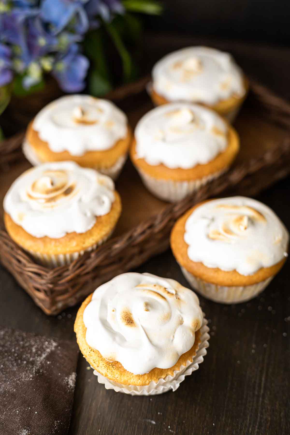 Six cupcakes topped with swirled white frosting are arranged in a wicker tray, with one additional cupcake in front on a dark wooden surface. Flowers are in the background.