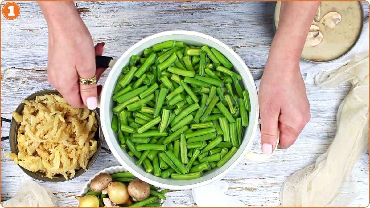 A person holds a pot filled with cut green beans on a wooden table, surrounded by chopped potatoes, whole potatoes, mushrooms, and a bowl of soup.