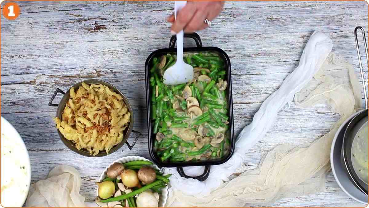 A hand stirs a casserole dish filled with green beans and mushrooms; cooked noodles, vegetables, and kitchen utensils are on a rustic white table.