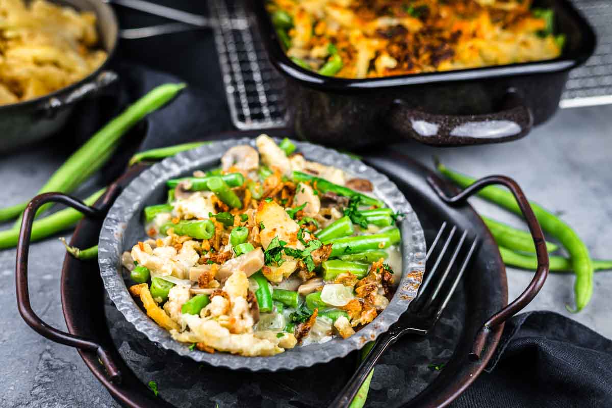 A serving of green bean casserole with crispy topping on a plate, with a fork beside it and a casserole dish in the background. Fresh green beans are scattered nearby.