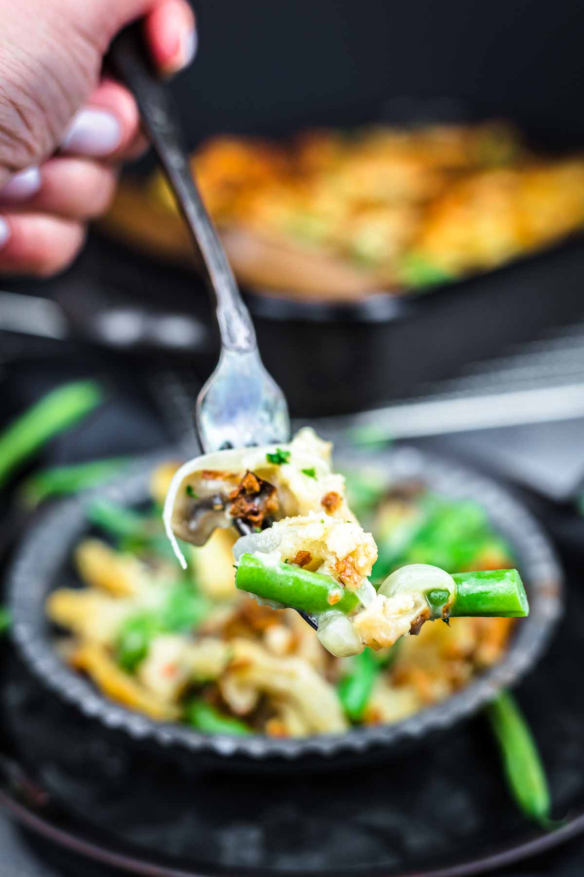 A close-up of a fork holding a bite of green bean casserole with mushrooms and crispy topping over a bowl.