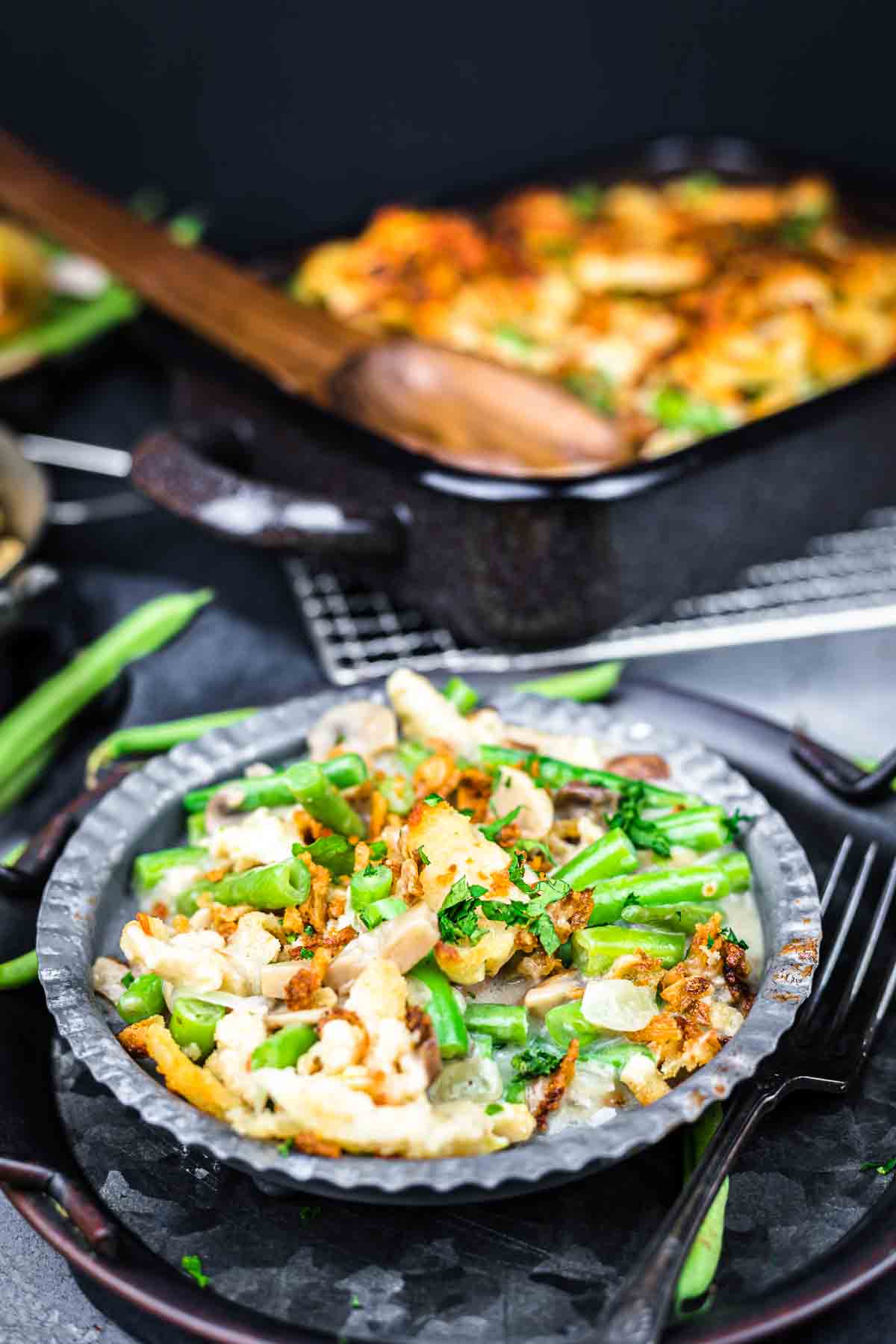 A plate of green bean casserole topped with crispy fried onions, garnished with herbs; a casserole dish and wooden spoon are in the background.