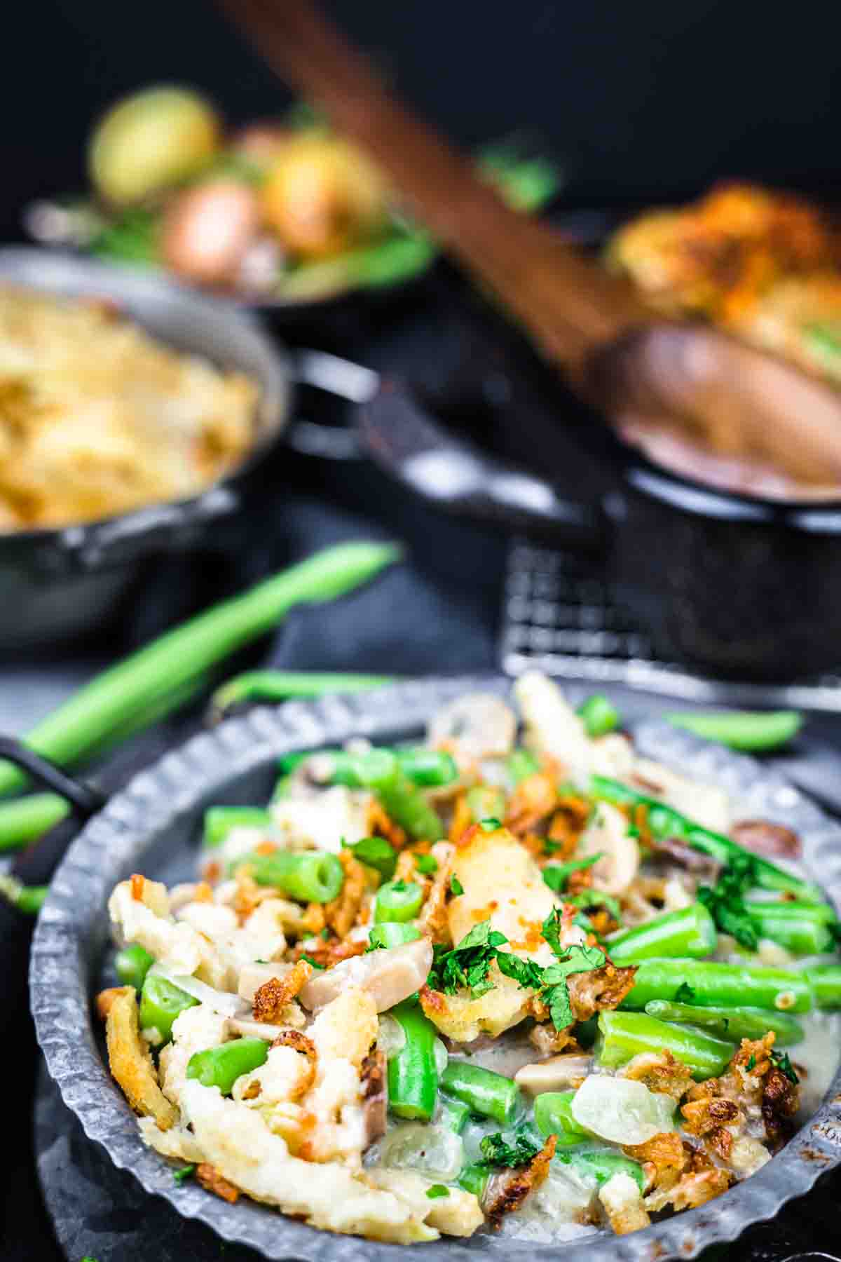 A metal plate filled with green bean casserole topped with fried onions and herbs, with pots and a wooden spoon in the background.