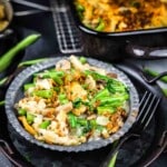 A close-up of a serving of green bean casserole with mushrooms and crispy onions on a dark plate, with a fork and casserole dish in the background.