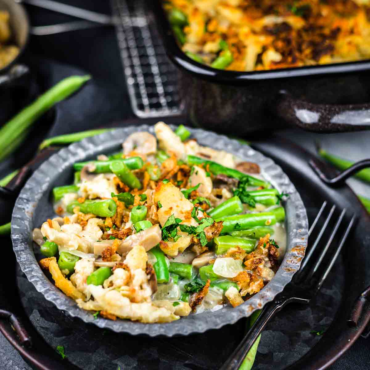 A close-up of a serving of green bean casserole with mushrooms and crispy onions on a dark plate, with a fork and casserole dish in the background.