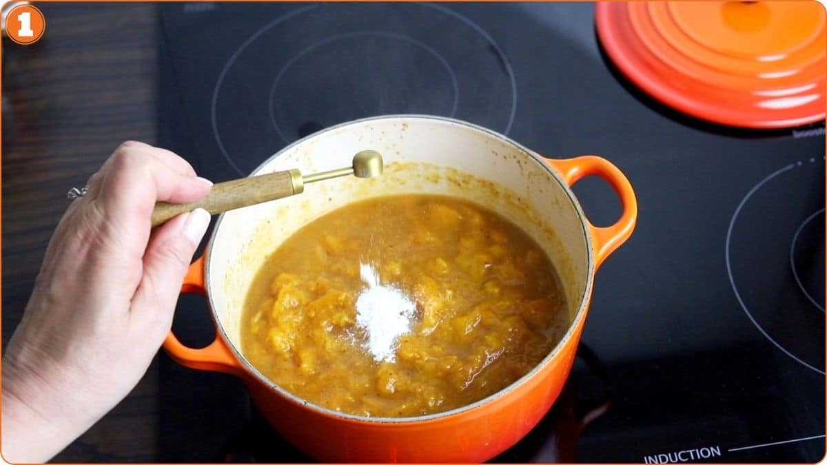 A hand adds a spoonful of white powder to an orange pot with simmering yellow stew on a black induction cooktop.