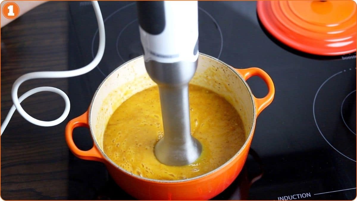 An immersion blender is blending soup in an orange pot on a black induction stovetop.