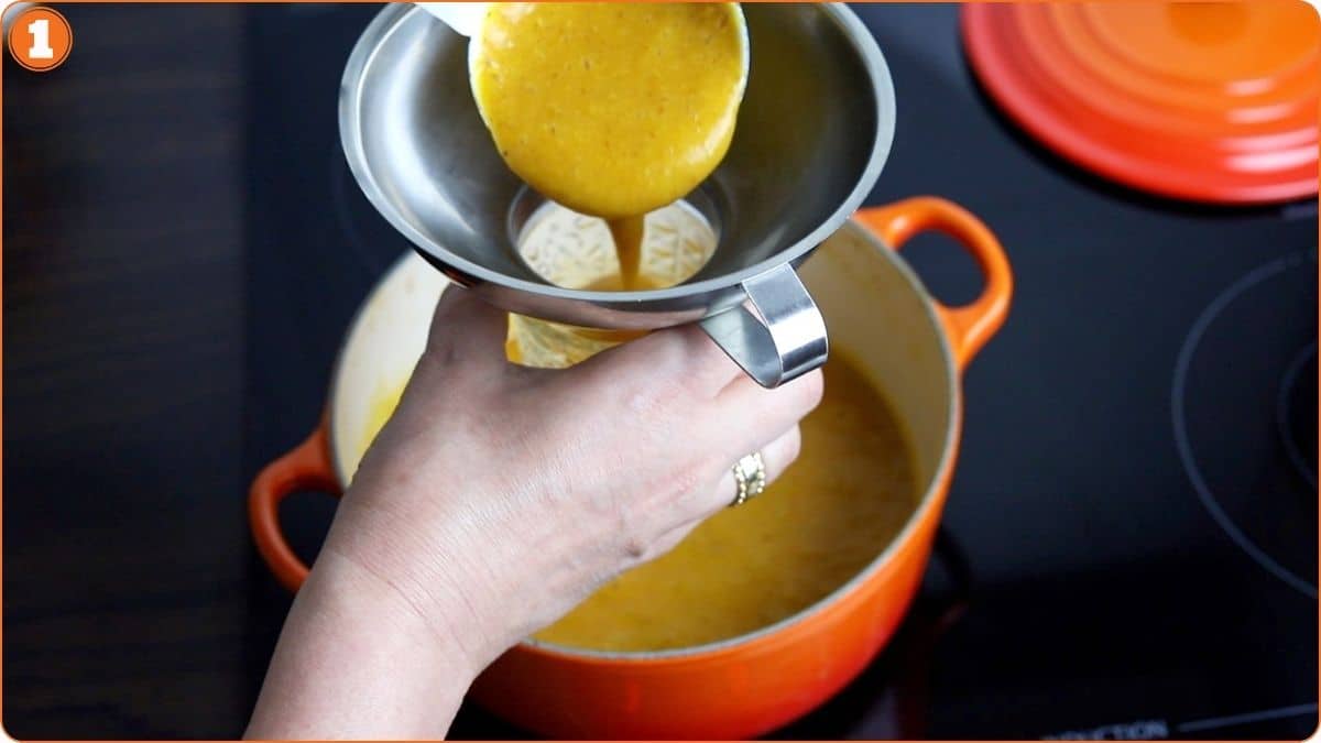 A person pours soup from a ladle through a funnel into a jar above an orange pot on a stovetop.