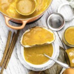 A slice of bread on a white plate is spread with orange pumpkin butter. A pot, jars of pumpkin butter, a spoon, and cinnamon sticks are nearby on a white wooden surface.