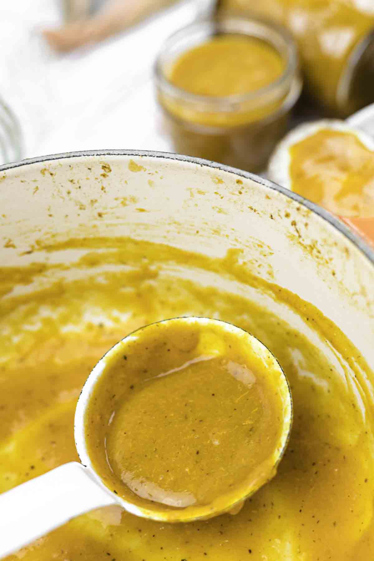A white ladle filled with thick orange-brown sauce or soup, held over a large pot with the same mixture; jars of the sauce are visible in the background.