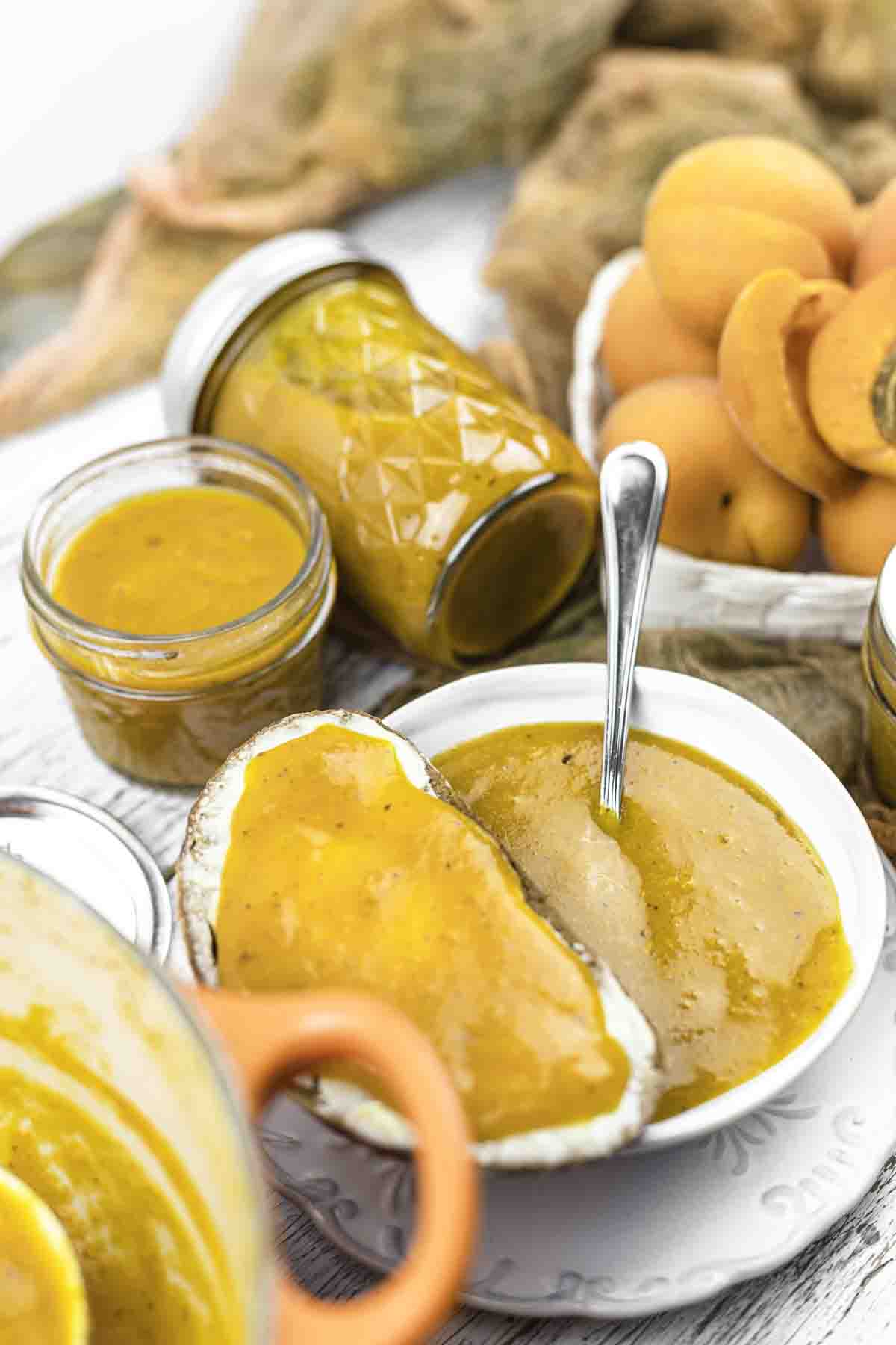 Jars and a bowl of orange apricot jam, with a slice of bread spread with jam, fresh apricots, and sweet potatoes in the background.