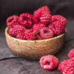 A wooden bowl filled with fresh raspberries, with a few raspberries scattered on a dark surface beside it.