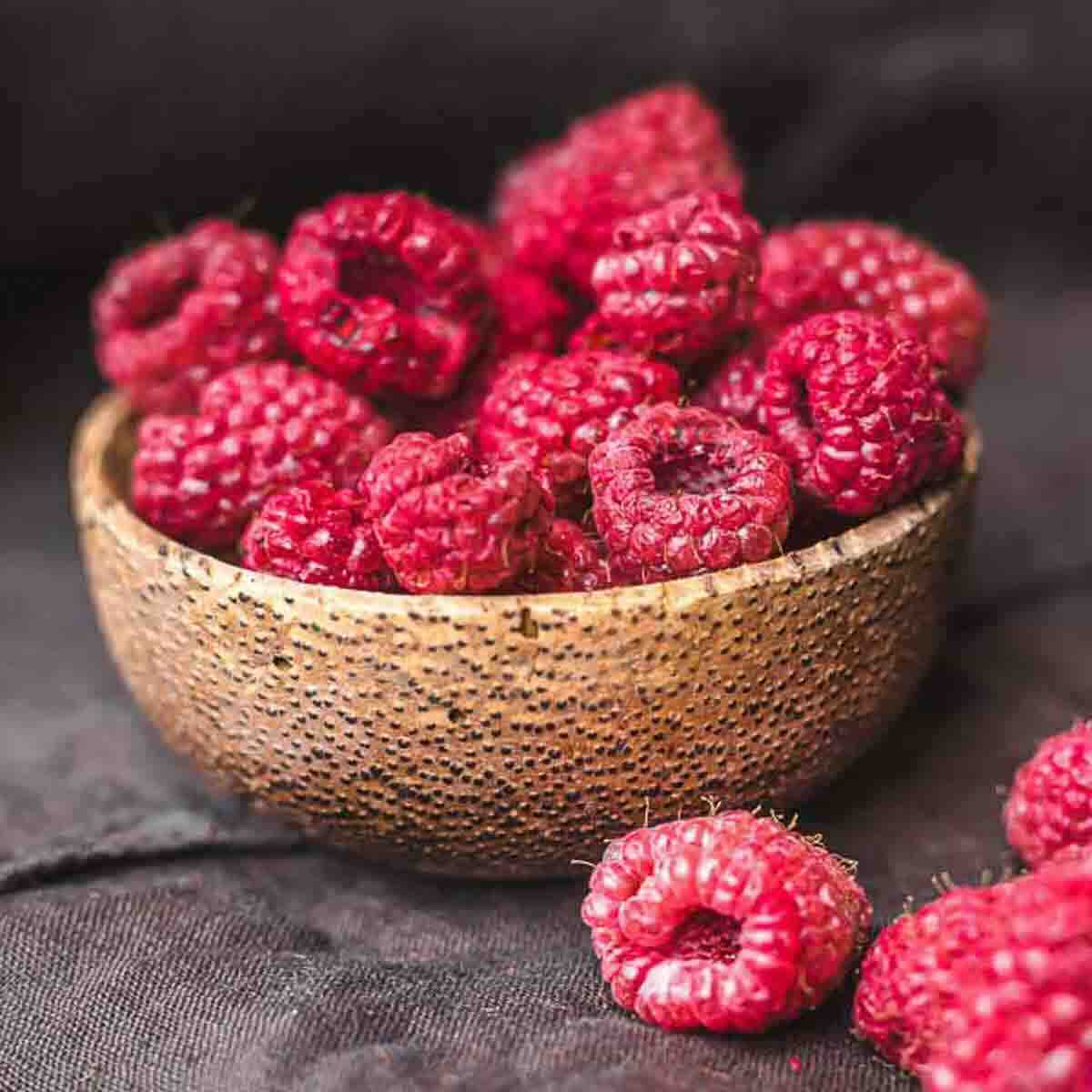 A wooden bowl filled with fresh raspberries, with a few raspberries scattered on a dark surface beside it.