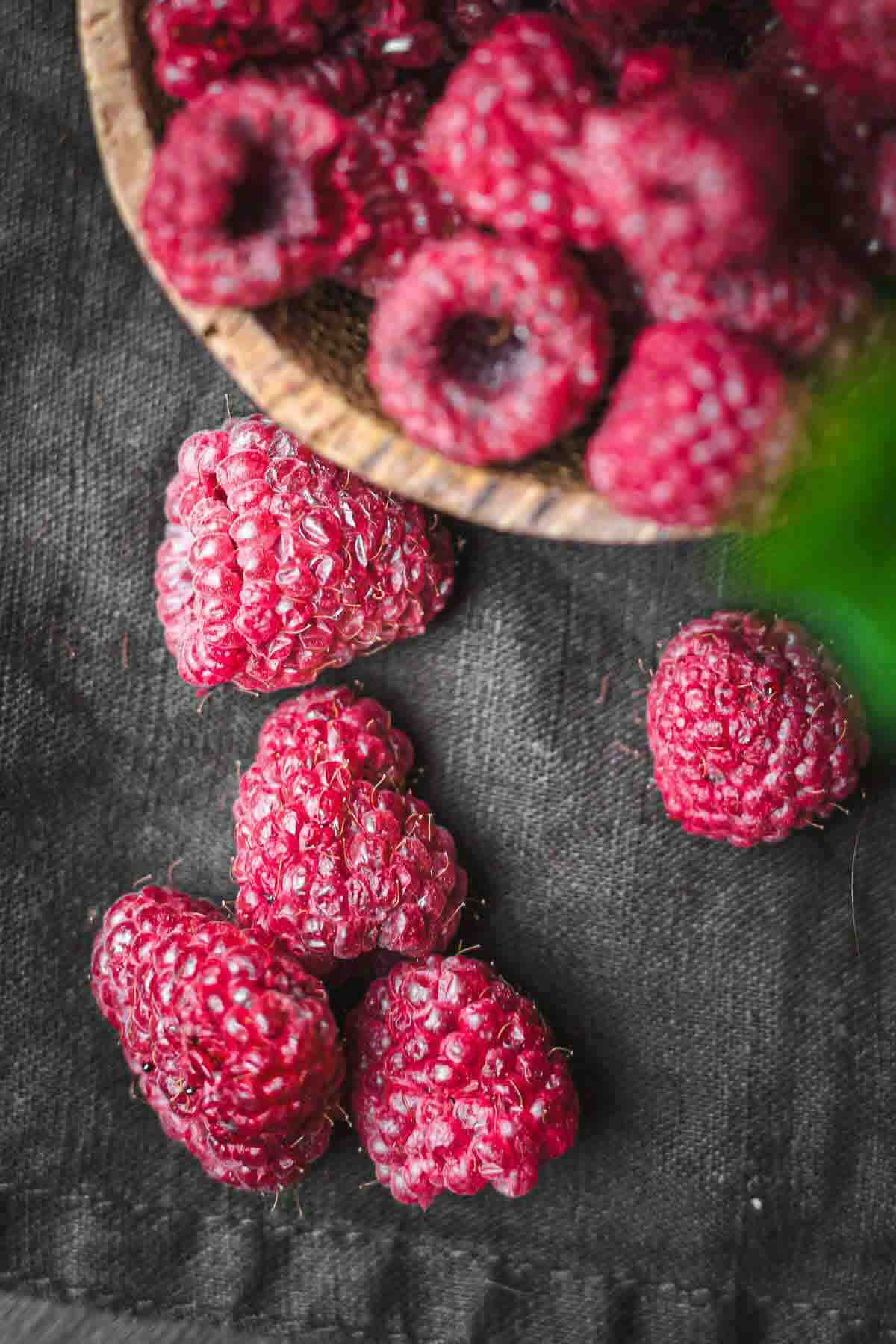 Several fresh raspberries are scattered on a dark fabric surface, with more raspberries in a wooden bowl above.