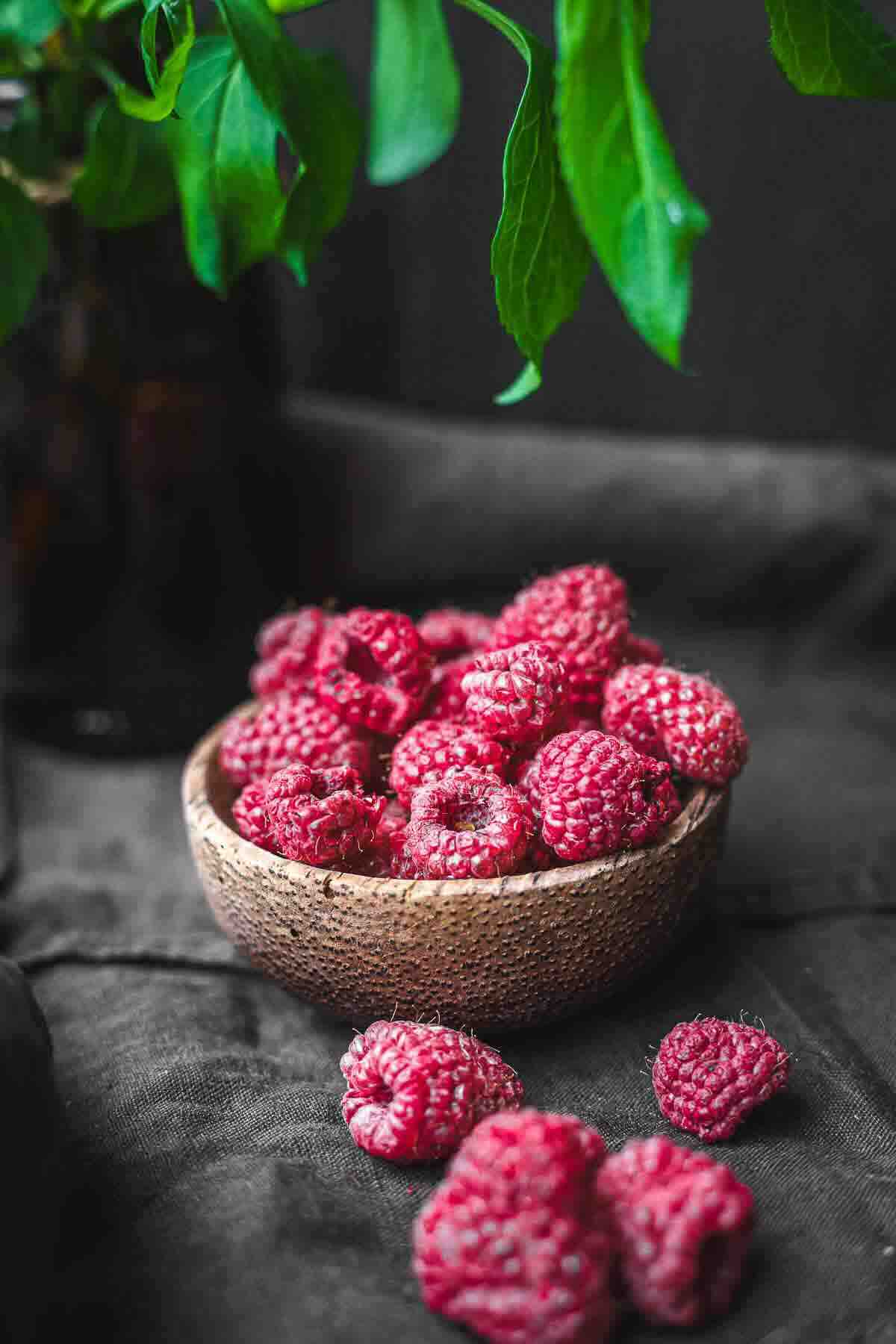 A wooden bowl filled with ripe raspberries sits on a dark cloth, with a few raspberries scattered in front and green leaves hanging above.