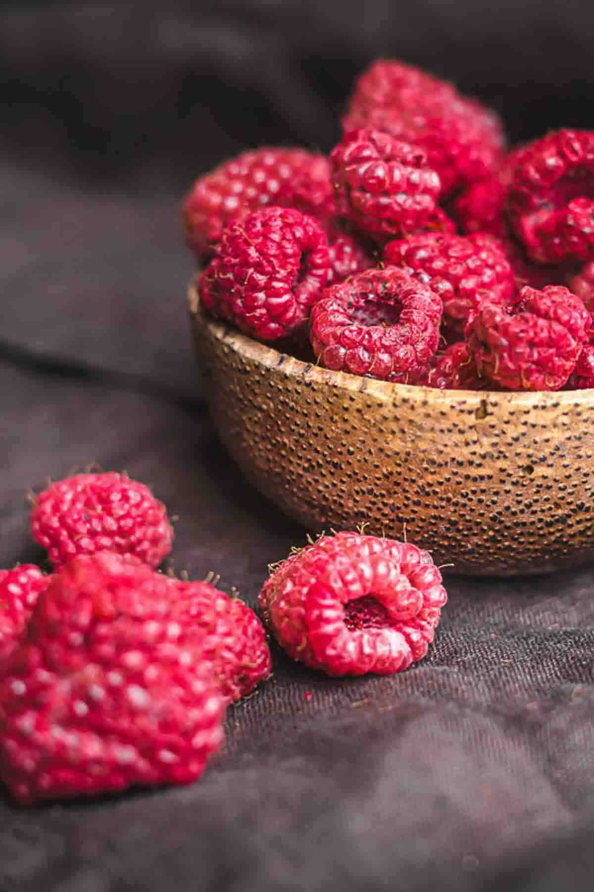 A textured bowl filled with fresh raspberries sits on a dark surface, with a few loose raspberries placed beside the bowl.