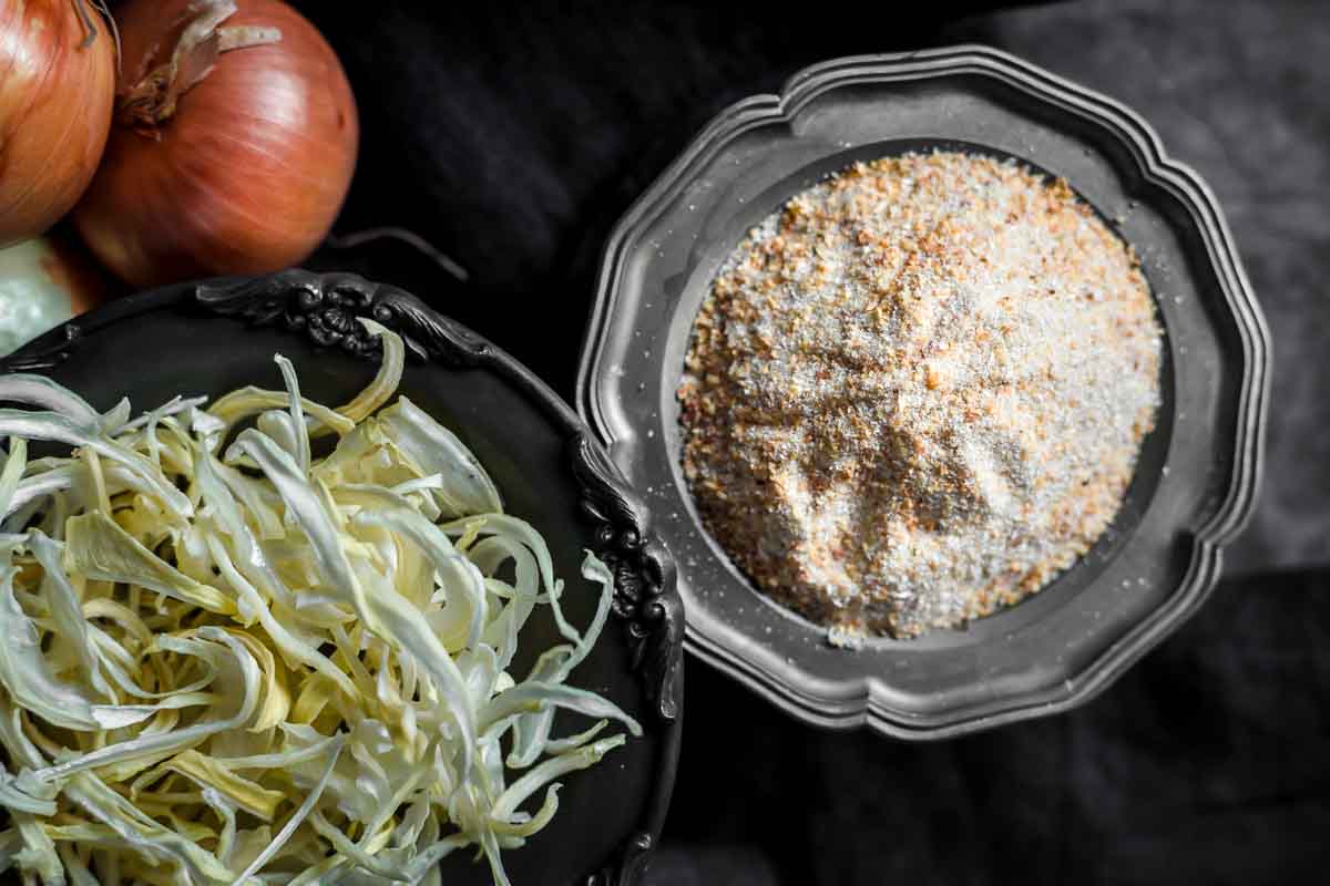 A bowl of dried, shredded onions next to a bowl of onion powder, with whole onions in the background on a dark surface.