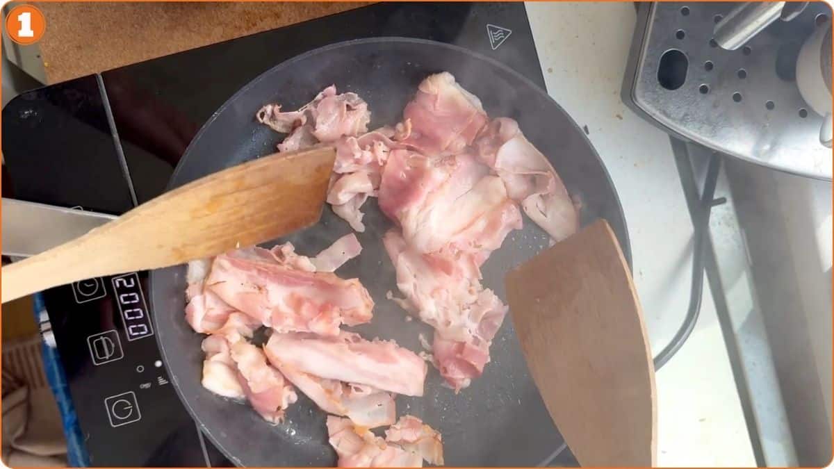 Raw pieces of meat are being stirred in a frying pan with two wooden spatulas on a stovetop.