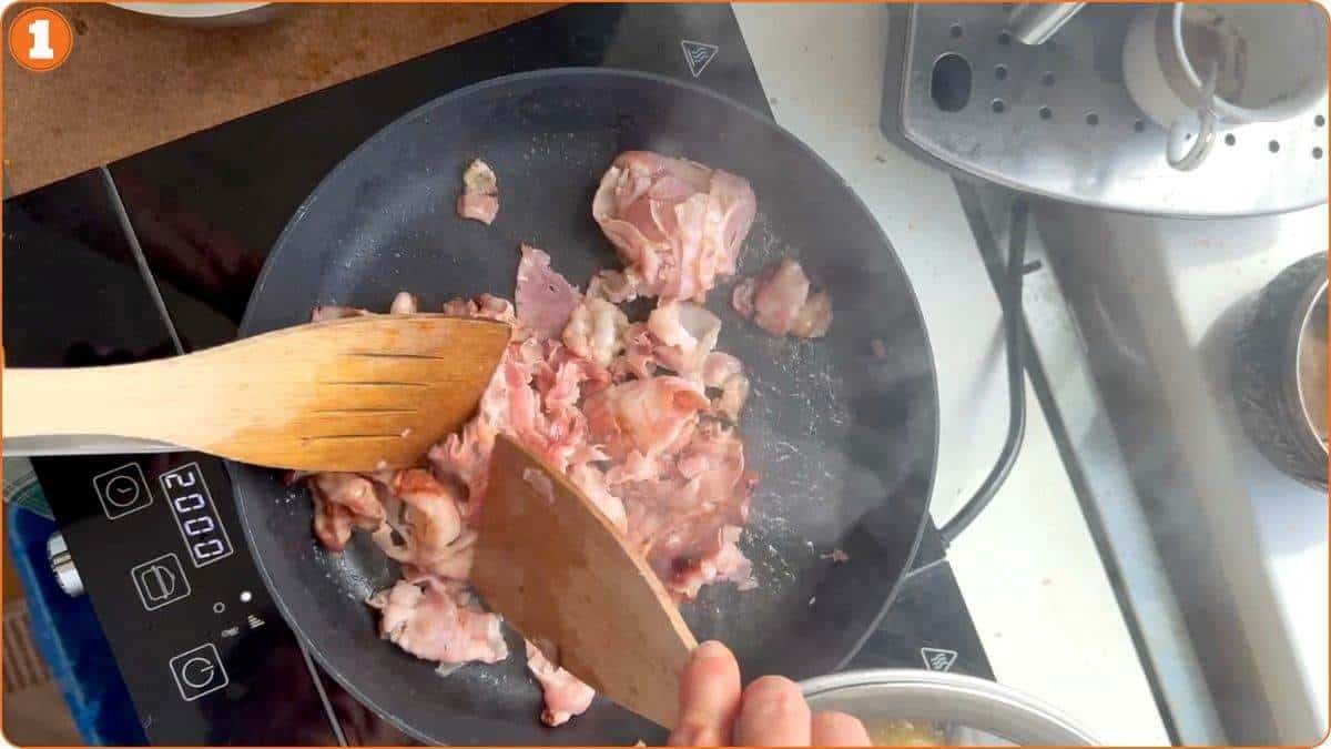 Raw chicken pieces being stirred with wooden utensils in a frying pan on an electric stovetop.