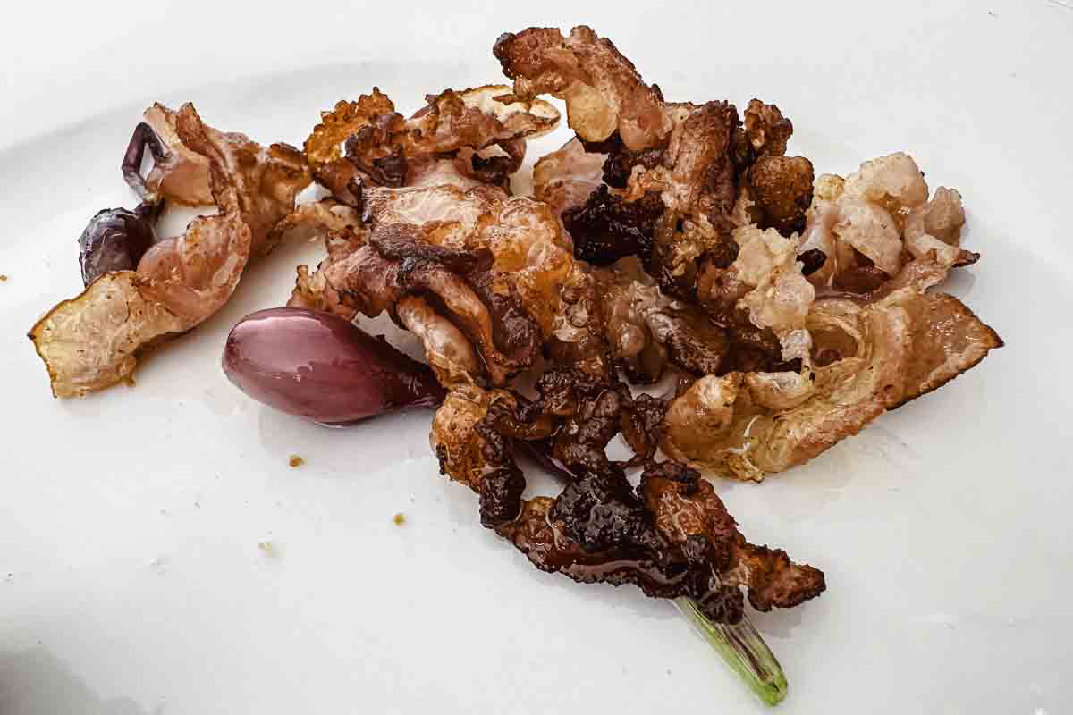 Crispy cooked bacon pieces and a few small red onions on a white plate.