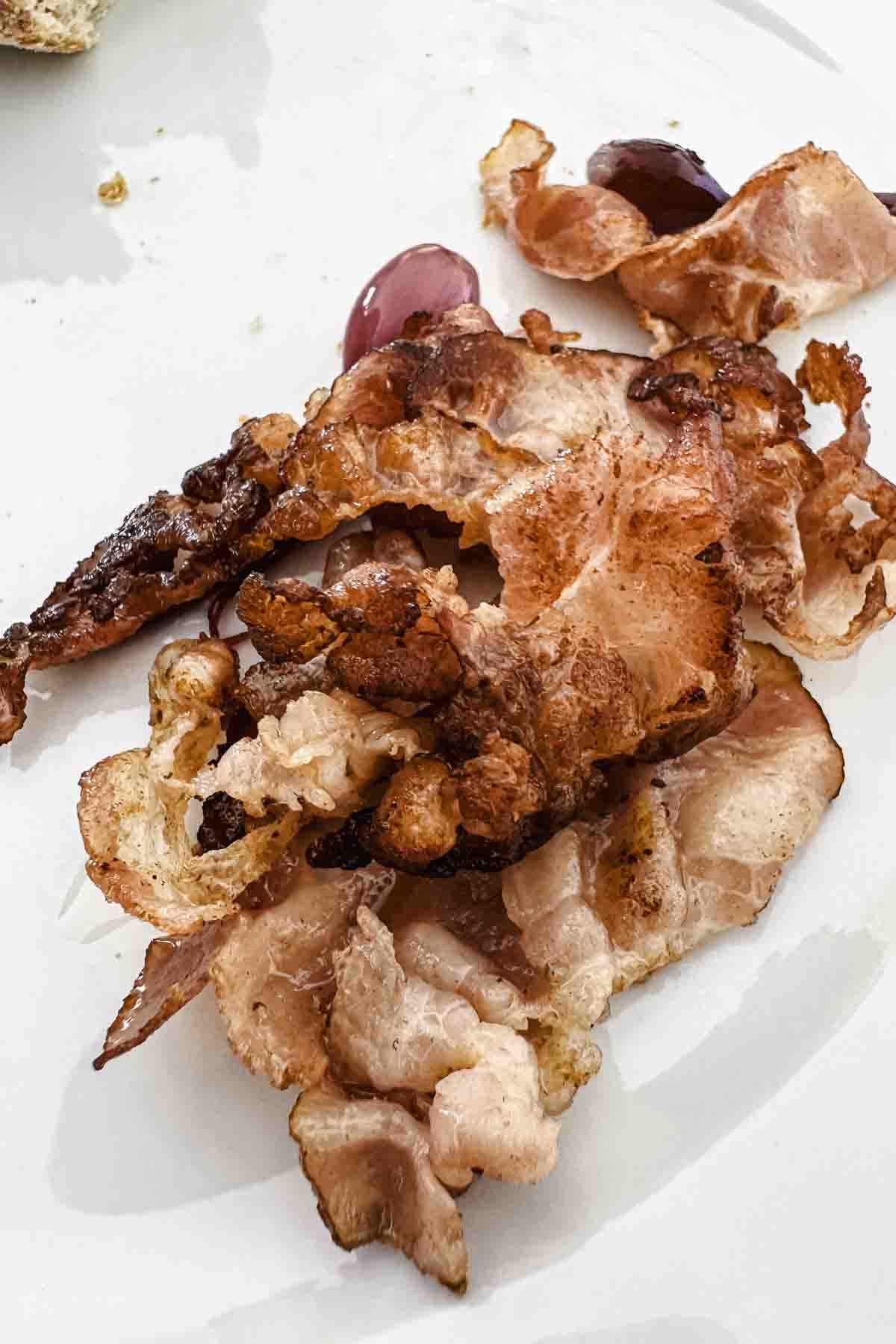 Crispy cooked bacon strips on a white plate with a few small purple grapes in the background.