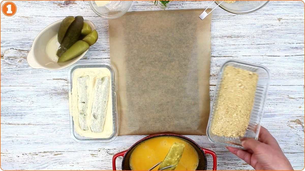 A table with breaded fish fillets, whole pickles, a bowl of egg wash, and a container of breadcrumbs arranged around a sheet of parchment paper.