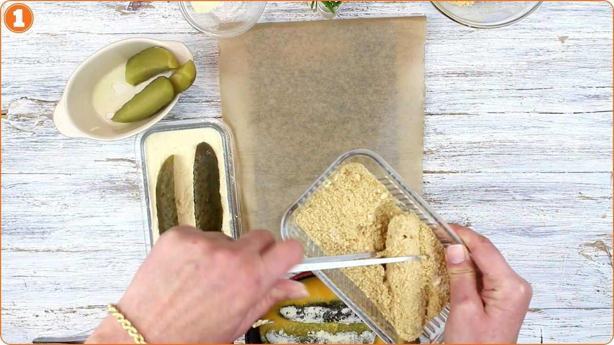 A person slices breaded food over parchment paper, with pickles and other ingredients nearby on a white wooden surface.