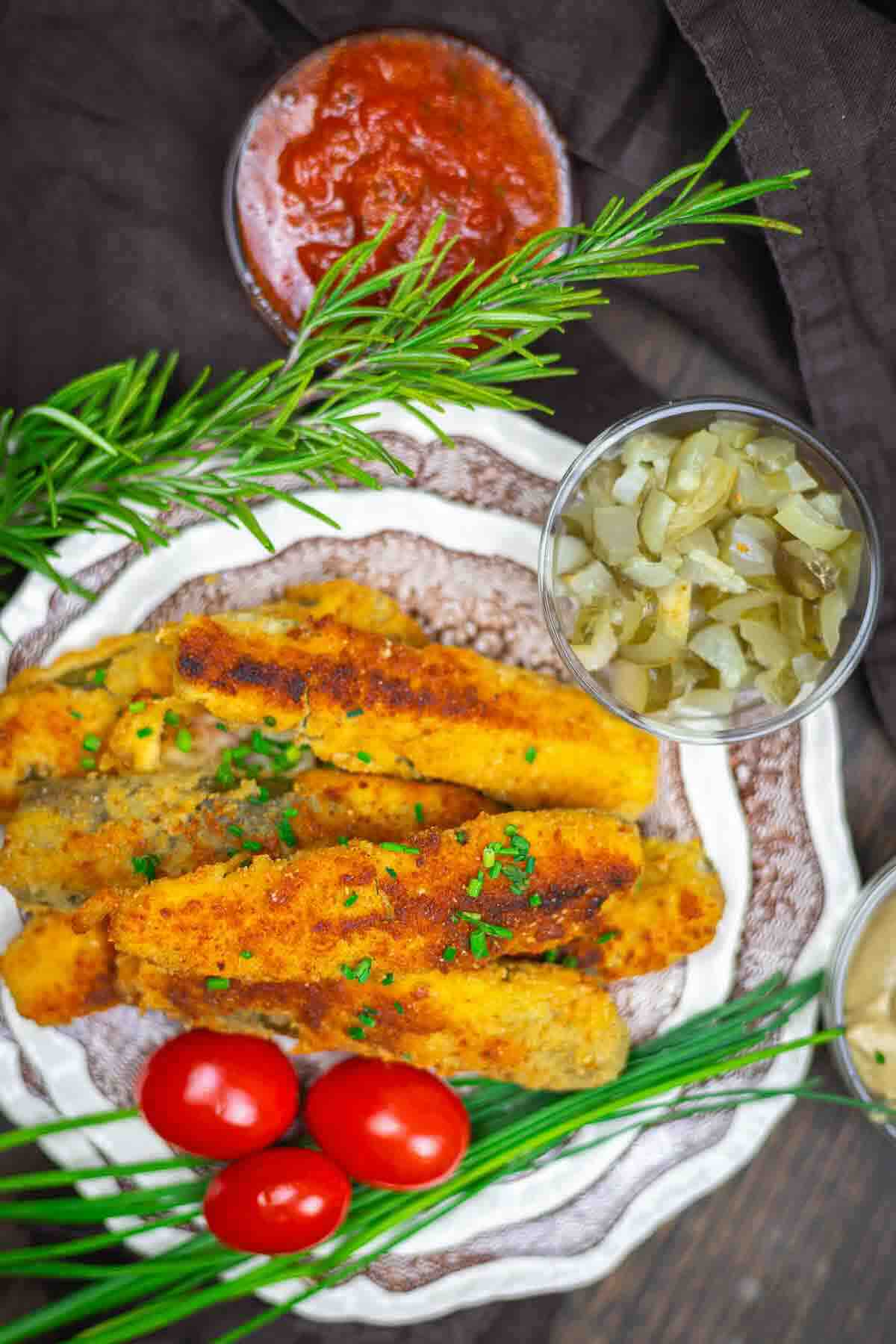 A plate of breaded and fried zucchini sticks garnished with herbs, served with cherry tomatoes, chopped pickles, and dipping sauces.
